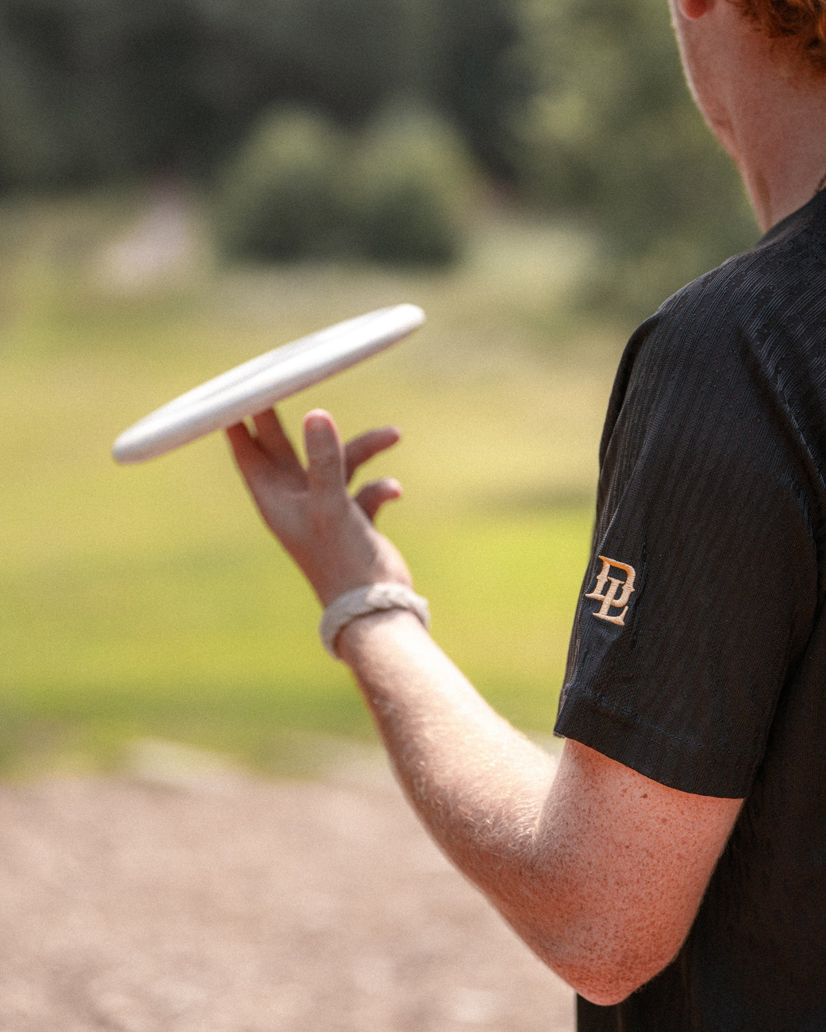 A person outdoors holding a white disc near their hand, wearing a black short-sleeve athletic shirt with a small gold logo on the sleeve.